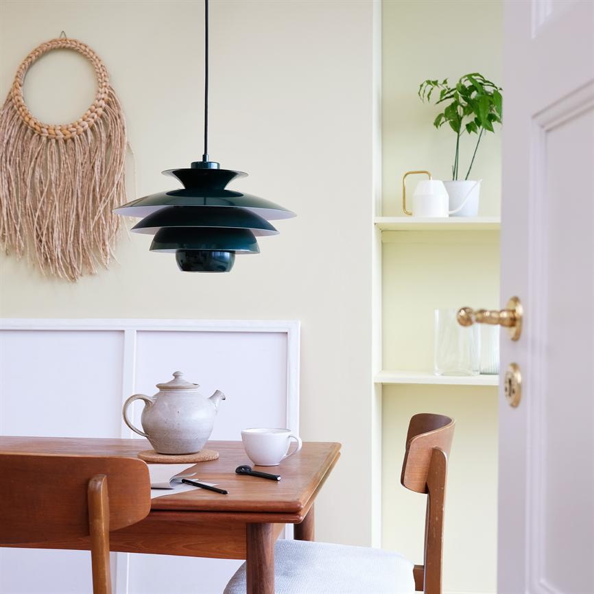 Sort lagdelt pendel hænger over et træbord med tekande og kop; i en lys, minimalistisk spisekrog med vævet vægophæng, hylder og en grøn plante.