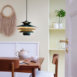 Pendellampe hænger og kaster varm belysning over et træbord; et tekande, en kop, notesbog og pen står på bordet i et hyggeligt spisehjørne med macramé, hylde og åben dør.