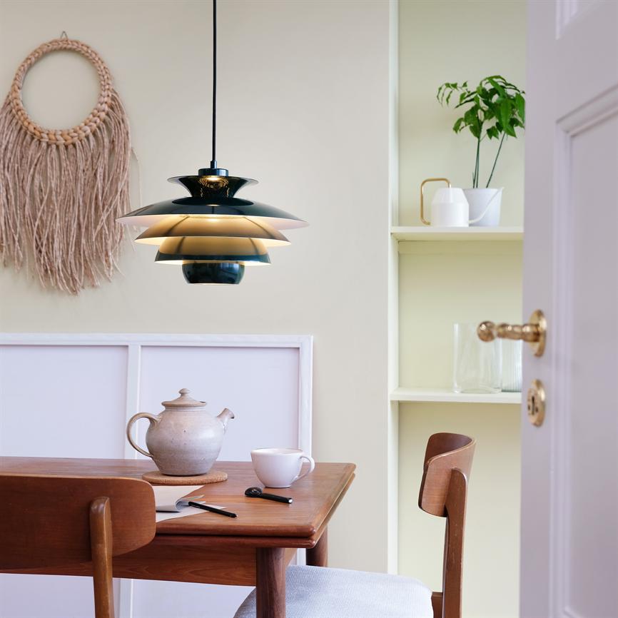 Pendellampe hænger og kaster varm belysning over et træbord; et tekande, en kop, notesbog og pen står på bordet i et hyggeligt spisehjørne med macramé, hylde og åben dør.