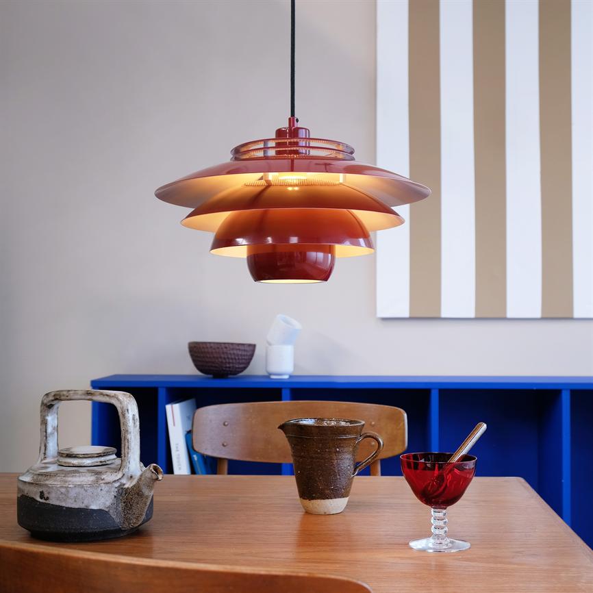 En rød flerlags hængelampe hænger og kaster varm belysning over et træbord med tekande, keramikkrus og rød glasskål; bagved en blå reol og lodrette beige-hvide striber på væggen.
