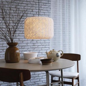 En vævet pendelampe hænger og kaster varmt lys over et rundt spisebord med vaser, skåle og tekande, flankeret af træstole foran hvidmalet murstensvæg og tynde gardiner.