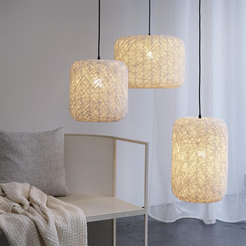 Tre hængende, trådflettede lampeskærme lyser blødt, svævende over en lys, fletrygget stol med pude og vaffelvævet tæppe foran et gennemsigtigt hvidt gardin og mørkt betongulv.