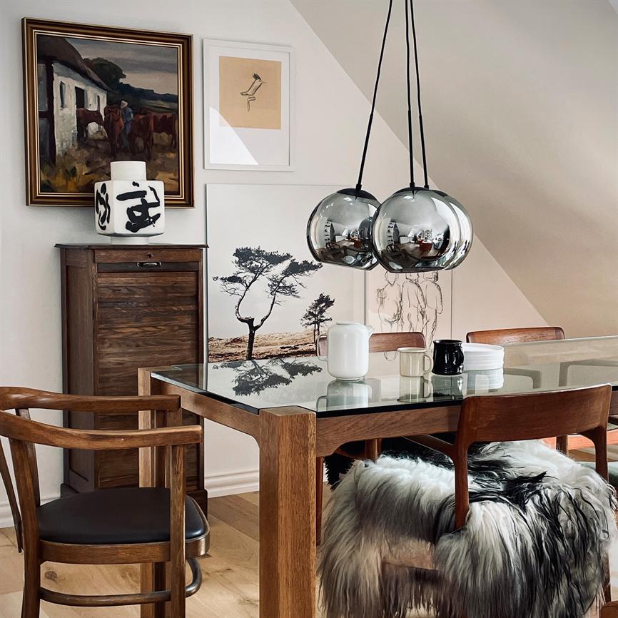 Spisebord i træ med glasplade står midt i et loftshjørne, to blanke kuglependler hænger, kunst og skænk bagved, træstole med fårskind og keramik på bordet.