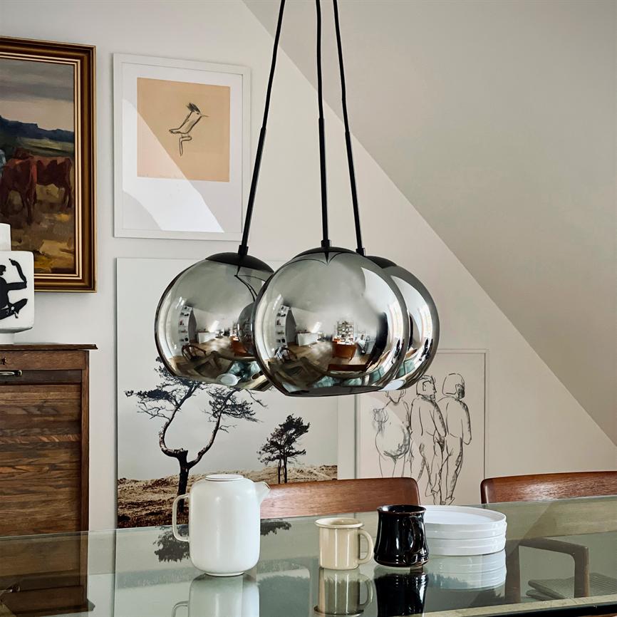 Tre spejlblanke kuglependler hænger og reflekterer rummet; over et glasbord med kande, krus og tallerkener; i en lys spisestue med flere kunstværker på væggen.