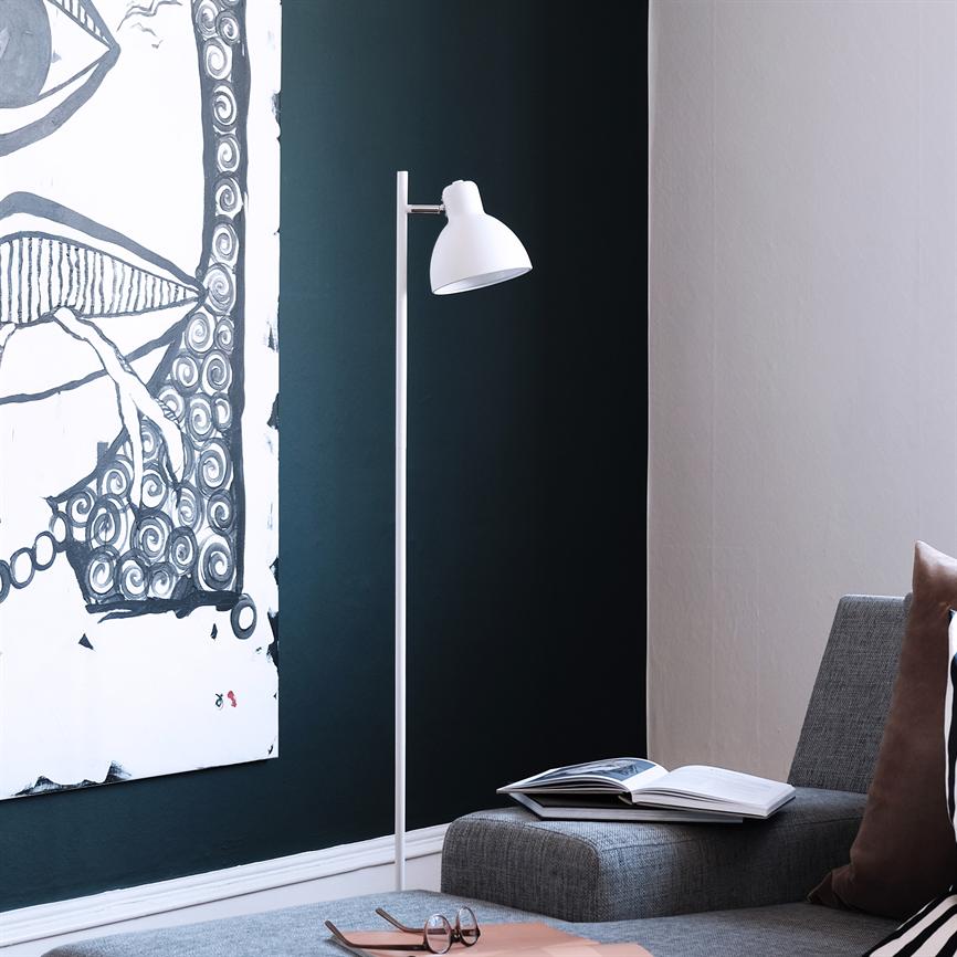 En hvid gulvlampe står ved en grå sofa og peger mod en mørk grøn væg; et sort-hvidt maleri hænger til venstre, bog og briller ligger på sofaen.
