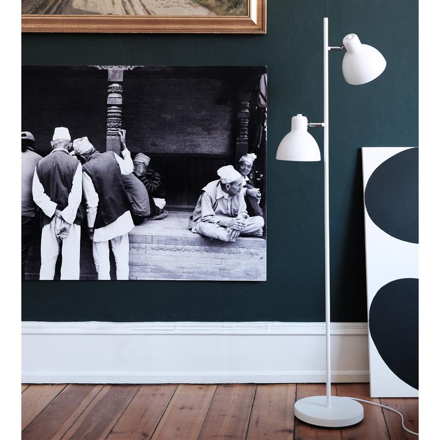 Stort sort-hvidt fotografi viser en gruppe ældre mænd i traditionelle huer, siddende og samtalende; ophængt på en mørkegrøn væg ved en hvid gulvlampe.