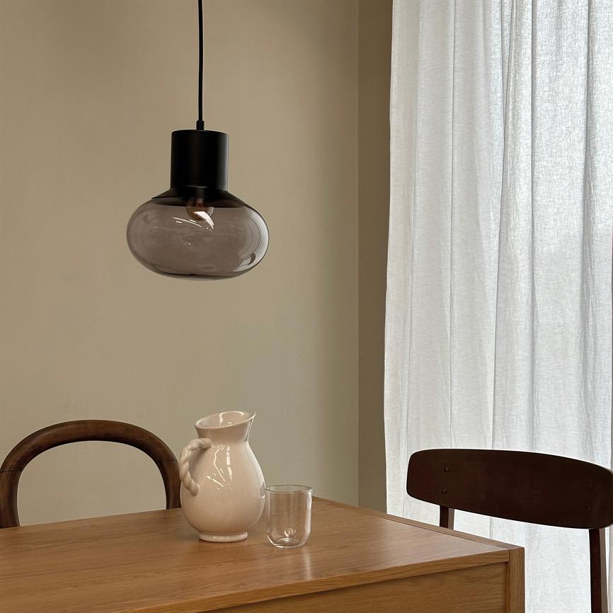 En mørk glaspendel hænger over et træbord; en hvid keramikkande og et klart glas står på bordet, med to træstole og lyse, gennemsigtige gardiner i baggrunden.