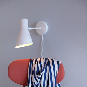 Væglampe lyser ned på en pink polstret stol, hvor et blåt-hvidt stribet tørklæde hænger afslappet over ryggen, mod en lys grå væg.