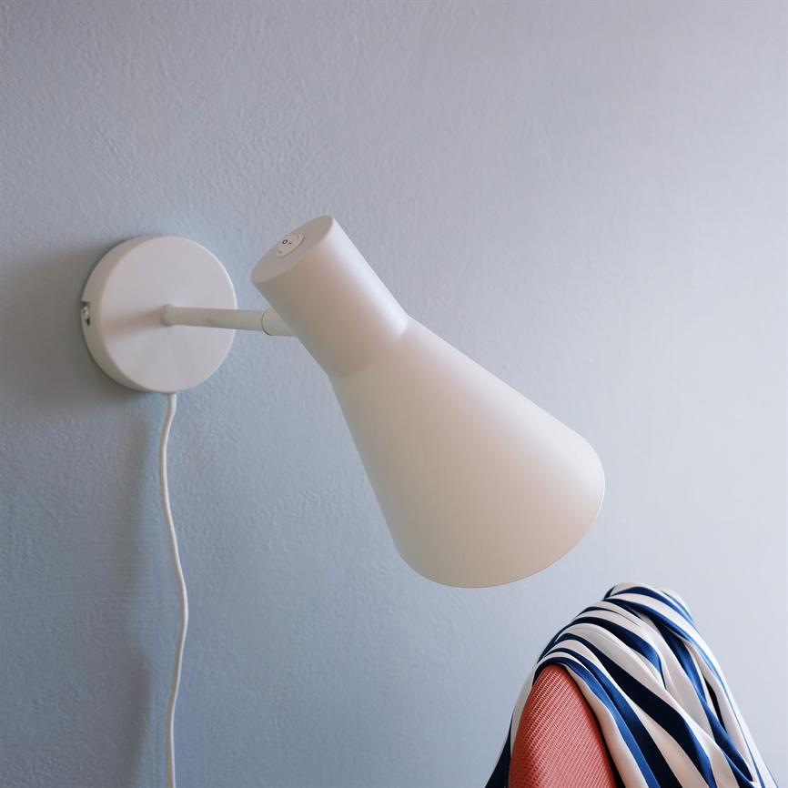 Hvid vægmonteret lampe peger ned mod en koralfarvet stol; et blå-hvidt stribet tørklæde hænger over stoleryggen, med en lysegrå væg og synlig ledning i baggrunden.