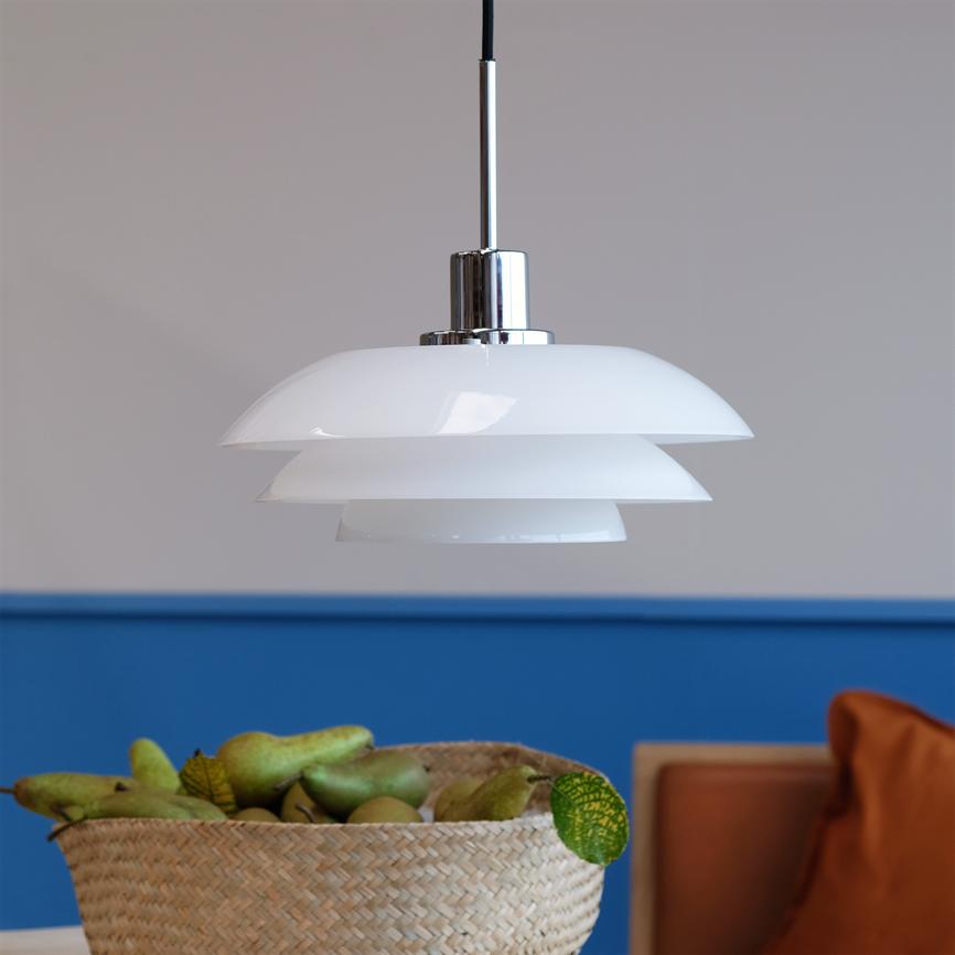 Hængelampe svæver over et bord; tre hvide, lagdelte skærme i blankt opalglas reflekterer lys. Bagved blå væg og kurv med pærer på bord, orange pude i hjørnet.