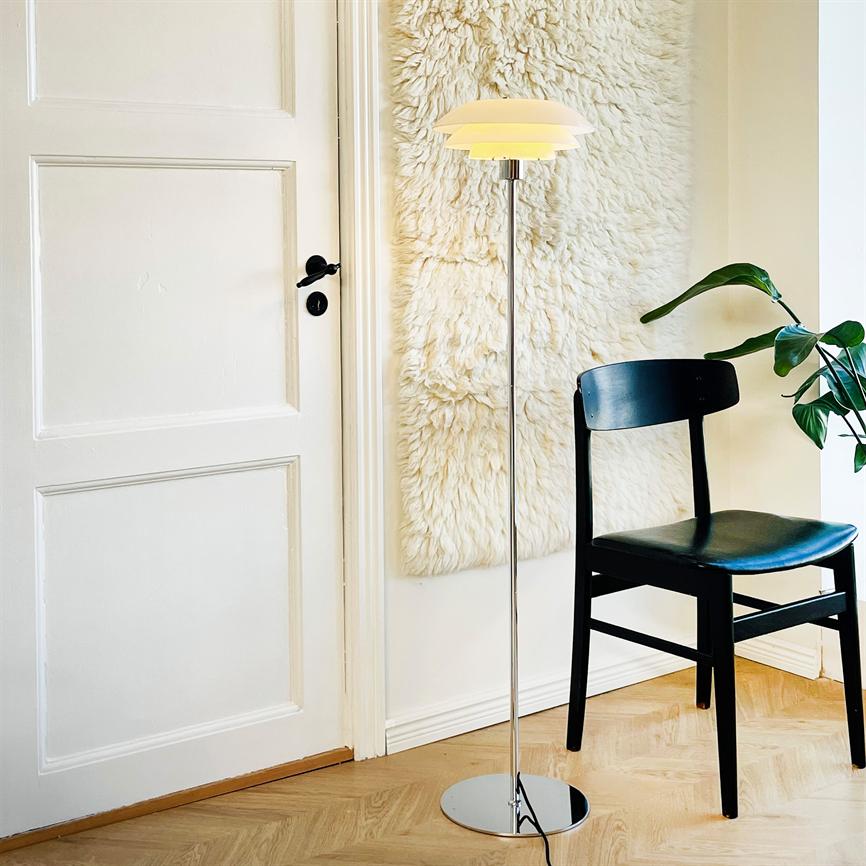 En slank krom gulvlampe står tændt foran et lyst, uldent vægskind; en sort træstol står ved siden af, foran en hvid dør og lyst parketgulv med en grøn stueplante.