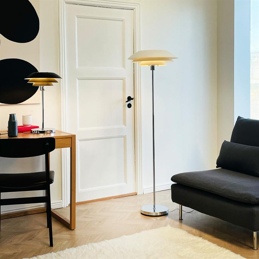 Gulvlampe står tændt ved en hvid dør i et lyst stuehjørne; skrivebordslampe lyser på et træbord med stol, mørk sofa og lyst tæppe i minimalistisk nordisk indretning.