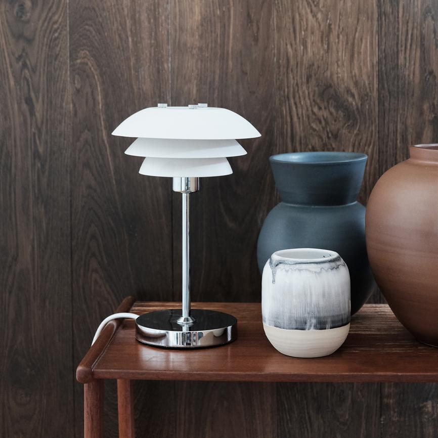 En hvid, flerlags bordlampe står på et mørkt træbord; den skinnende metalfod er flankeret af tre keramiske vaser i sort, hvid og terrakotta foran en mørk træpanelvæg.