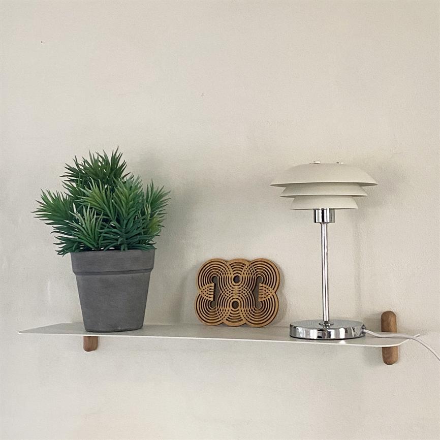 En grå potteplante står til venstre på en hvid væghylde; et træornament og en kromhvid bordlampe står ved siden af mod en lys væg.