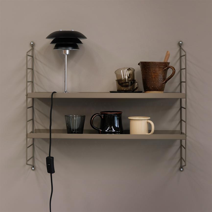 Væghylde viser en sort bordlampe, en brun lerkande med ske, to glas og tre krus; lampens ledning hænger ned. Kontekst: enkel beige væg.