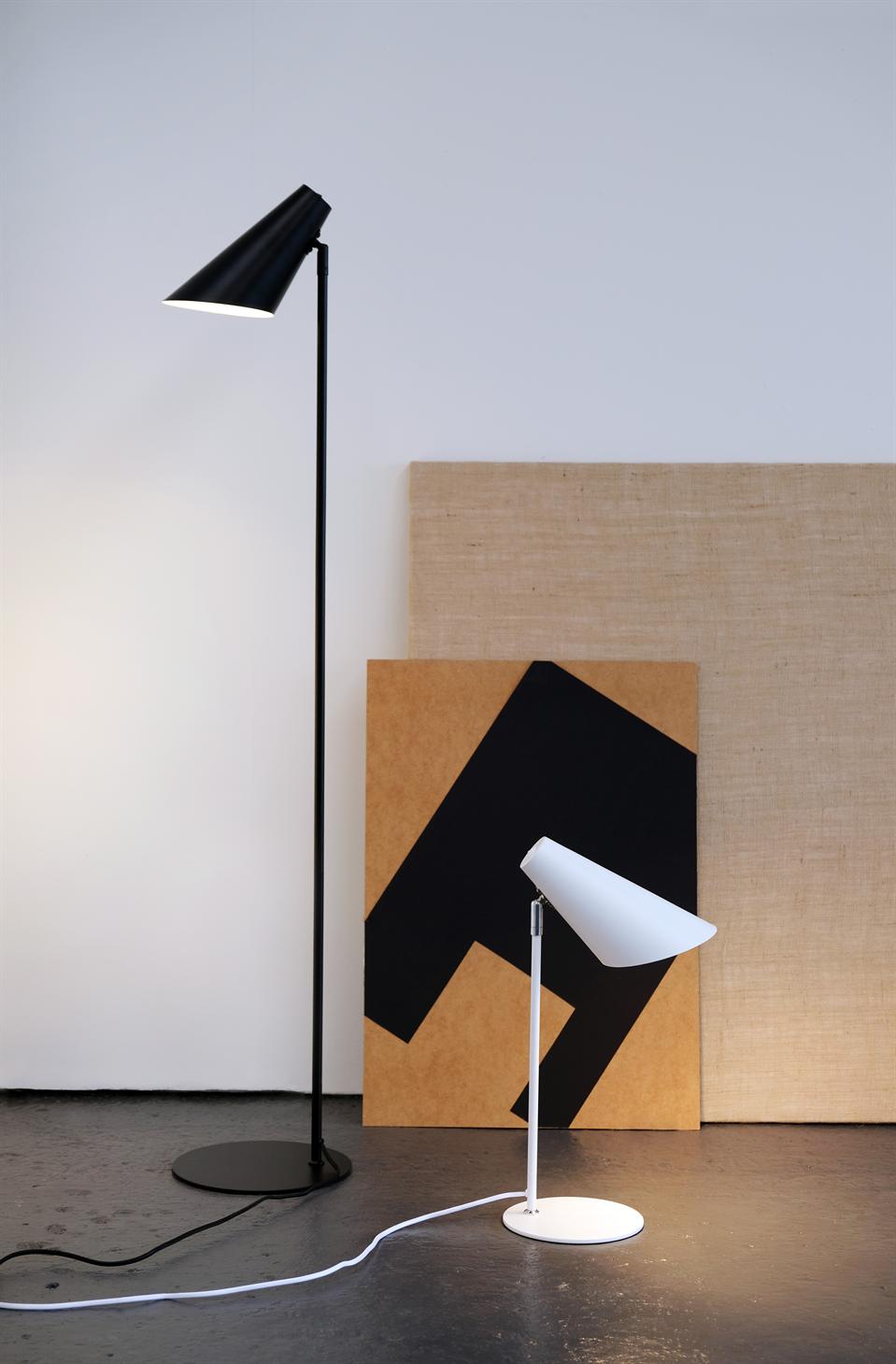 Sort gulvlampe og hvid bordlampe står tændt foran et geometrisk klistret træpanel og et naturfarvet lærred, placeret på mørkt betongulv i et lyst, minimalistisk rum.
