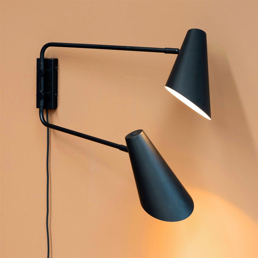 Sort væglampe med to justerbare kegleformede skærme, hvoraf den nederste kaster varmt lys nedad; lampen er monteret på en ferskenfarvet væg med synlig sort ledning.
