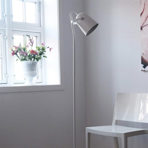 Gulvlampe står ved et vindue i et lyst, minimalistisk rum med en plaststol og en hvid vase med farverige blomster på vindueskarmen.