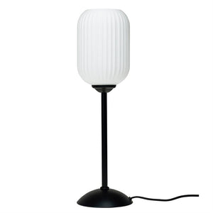 En bordlampe med ribbet, hvid cylindrisk skærm, står på en slank sort stang og rund sort fod med synlig ledning på hvid baggrund.