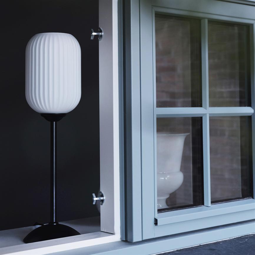 En hvid ribbet lampe står på en sort fod, placeret ved et åbent lyseblåt vindue med sprosser; mørk baggrund og en hvid vase ses gennem ruden.