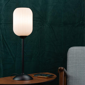 Bordlampe lyser blødt på et lille træbord med en smartphone; ved siden af en grå polstret stol foran en mørkegrøn, tekstureret baggrund.