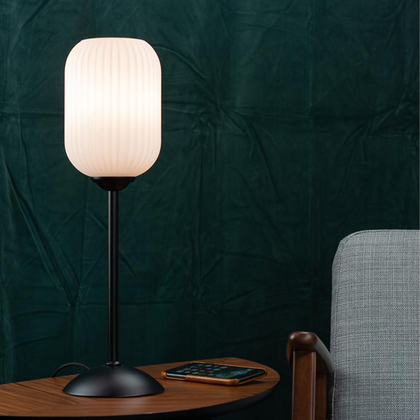 Bordlampe lyser blødt på et lille træbord med en smartphone; ved siden af en grå polstret stol foran en mørkegrøn, tekstureret baggrund.