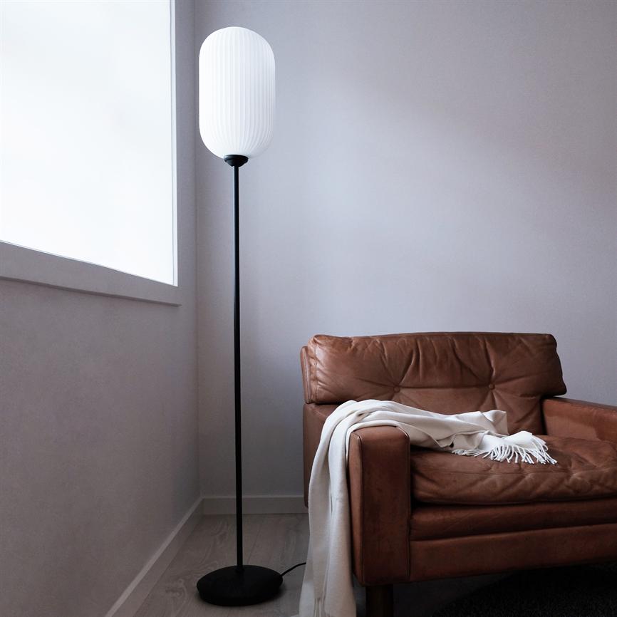 Brun læderlænestol står i et hjørne, et hvidt tæppe ligger over armlænet; ved siden af står en høj gulvlampe med hvid skærm, omgivet af lyst trægulv, grå vægge og et stort vindue.