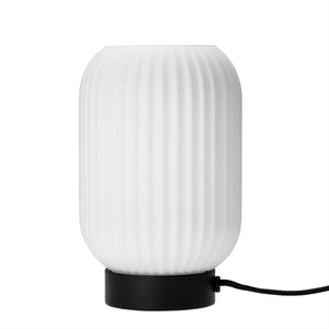 En hvid, lodret ribbet bordlampe står på en lav sort base; en kort sort stofledning rækker ud mod højre på en ensartet hvid baggrund.