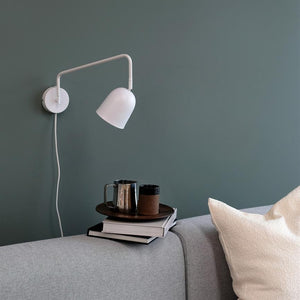 Hvid væglampe peger ned mod en bakke med bøger, et metalkrus og en termokop på armlænet af en grå sofa foran en grøn væg.