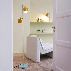 Stående lampe med gule skærme lyser ved siden af en lav lys loungestol; bagved hylder med pynt (kande, glas, bøger) i et roligt, lyst rum set gennem åben hvid dør.