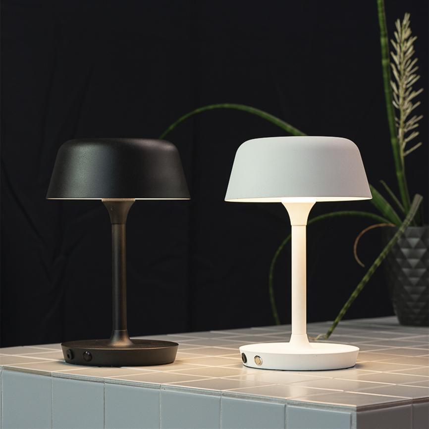 To bordlamper (en sort og en hvid) står tændte på et lysegråt flisebord; den hvide udsender varmt lys, baggrunden er mørk med grønne potteplanter.