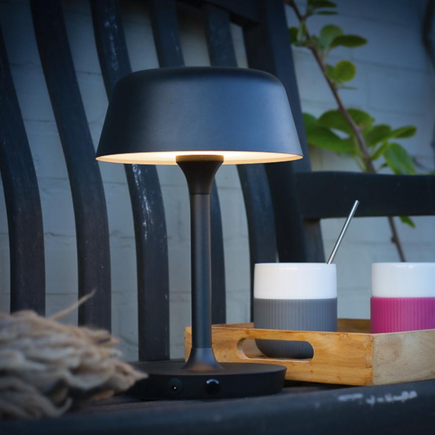 Bordlampe udsender blødt, varmt lys og står på et bord foran en sort træbænk; ved siden af ligger en træbakke med to krus (grå/hvid og pink/hvid) og en metalomrører.