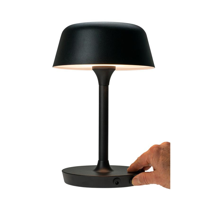 Sort bordlampe med rund skærm, der lyser nedad; en hånd drejer en knap på lampens flade bund; lampen står på et fladt underlag mod neutral hvid baggrund.