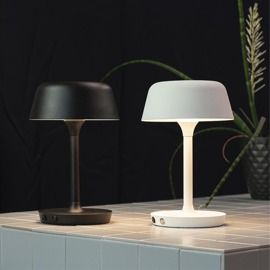 To bordlamper står på et flisebelagt bord; den ene sort slukket, den anden hvid og tændt; moderne, svampelignende design foran en mørk baggrund med en grøn plante.