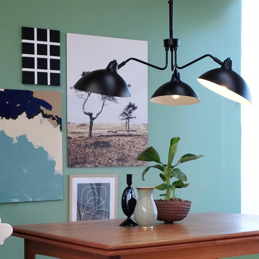 En sort, trearmet loftslampe hænger over et egetræsbord; den kaster varmt lys ned på vaser og en lille grøn stueplante, foran en lysegrøn væg med ophængte plakater.