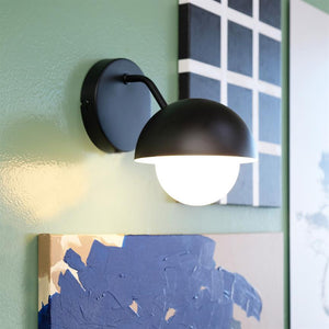 Væglampe — kaster blødt lys — monteret over farverige malerier på en lysegrøn væg, sort halvskærm med hvid rund pære.