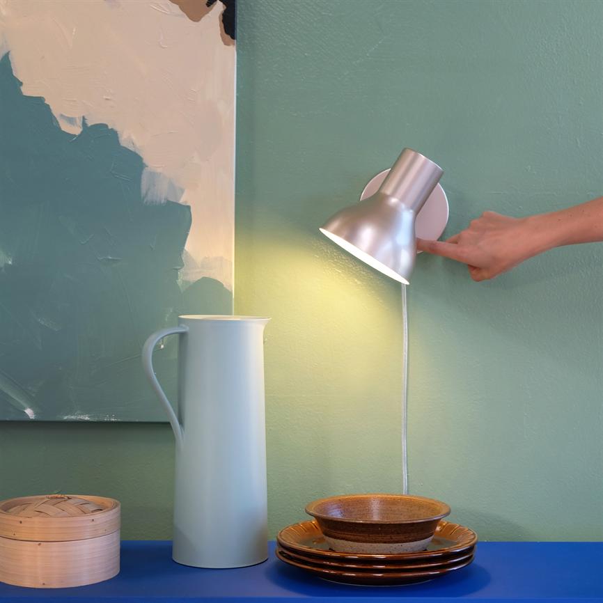 Væglampe tændes af en hånd mod en lysegrøn væg; lyset rammer en blå hylde med en høj hvid kande, stabel af brune skåle og et abstrakt maleri.