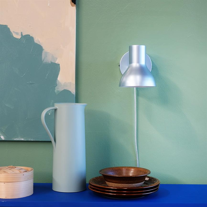 En lyseblå kande står på en blå hylde; en sølv væglampe lyser ned; brune tallerkener og en skål ligger ved siden af; grøn væg med delvist abstrakt maleri i baggrunden.