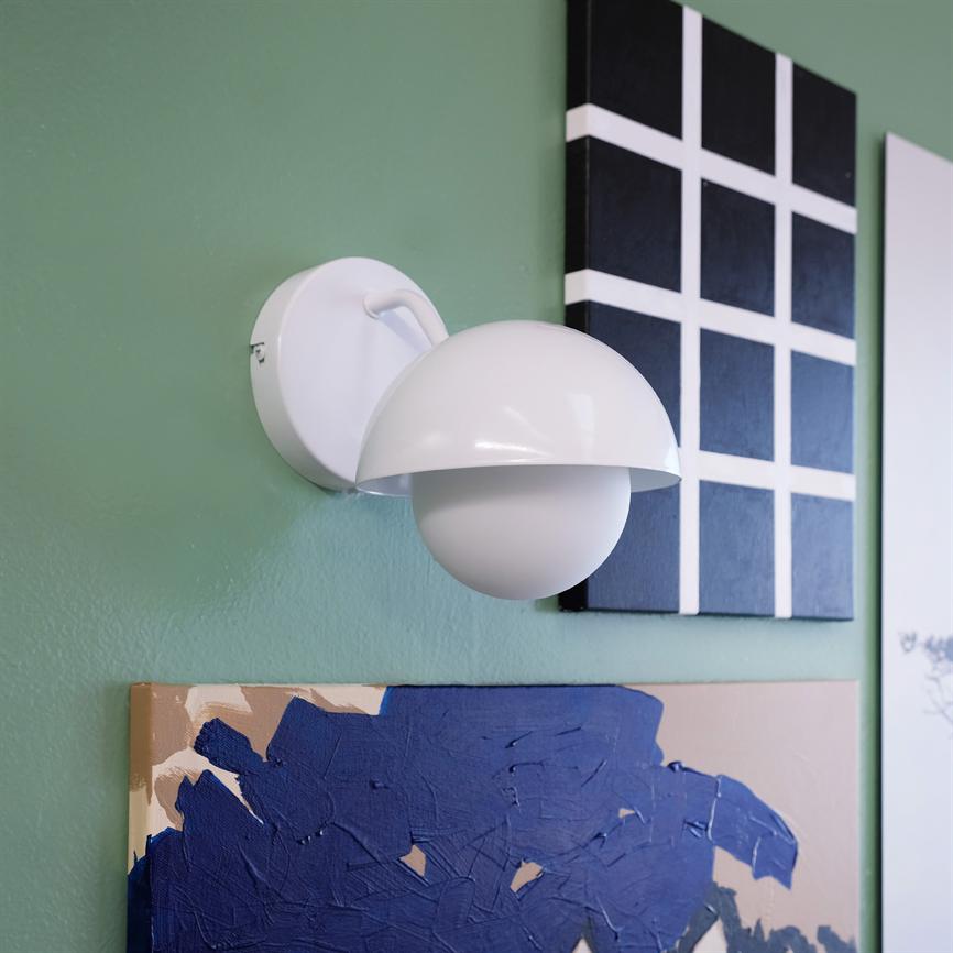 Hvid væglampe monteret på grøn væg, halvkugleformet skærm over rund pære; omgivet af tre malerier — sort-hvid ternet panel, blå tekstureret lærred og et lyst kunstværk.