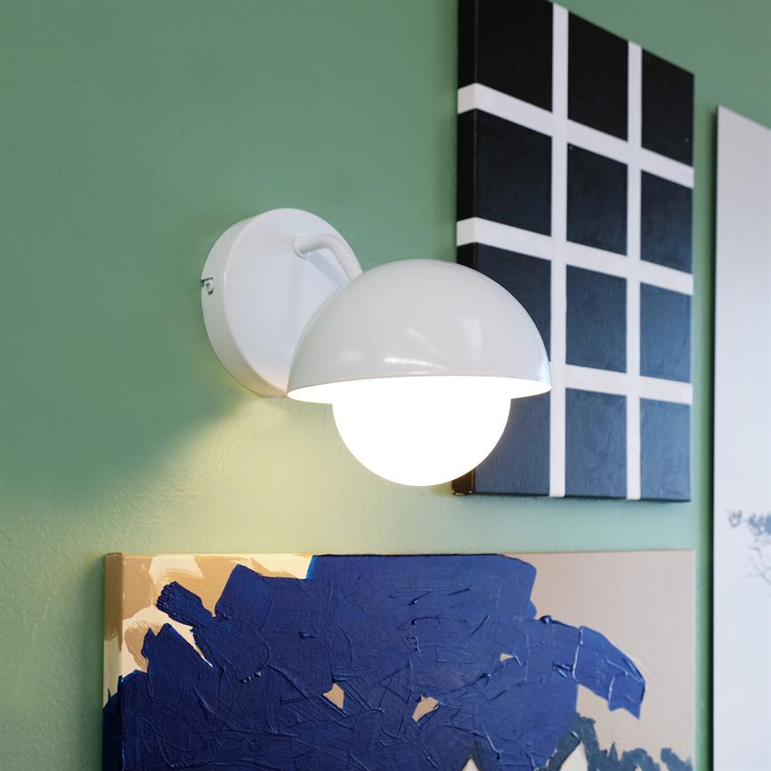 En hvid, halvkugleformet væglampe lyser klart; den er monteret på en grøn væg over to abstrakte malerier — et sort-hvidt gitter og et blåt, struktureret lærred.