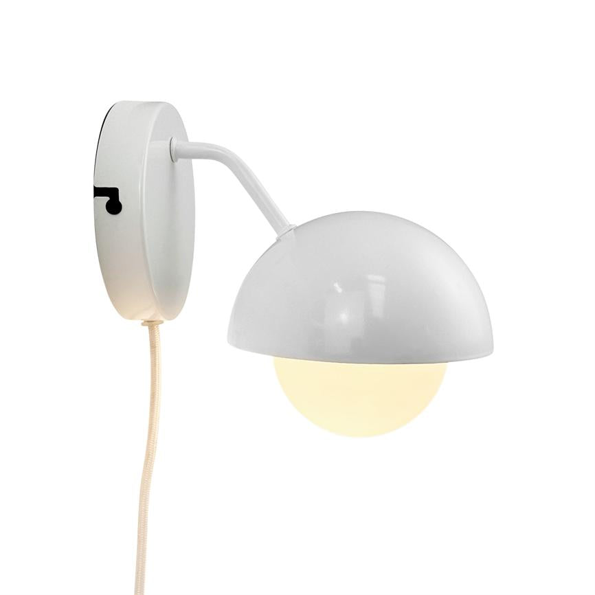 Væglampe: en hvid arm holder en halvkuppelformet skærm, som lyser en opal rund pære; monteret på rundt vægbeslag med tekstilledning mod hvid baggrund.
