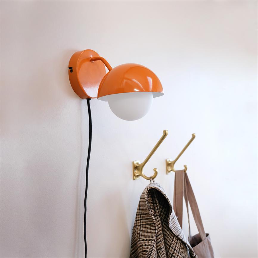 En orange væglampe hænger monteret på en hvid væg; dens runde skærm peger nedad; under den sidder to messingknager med en ternet frakke og en stoftaske.