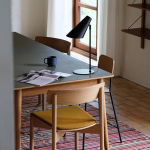 En sort bordlampe står på et grønt spisebord sammen med et åbent magasin og en blå kop; omgivet af træstole, et stribet tæppe og et vindue i naturligt dagslys.