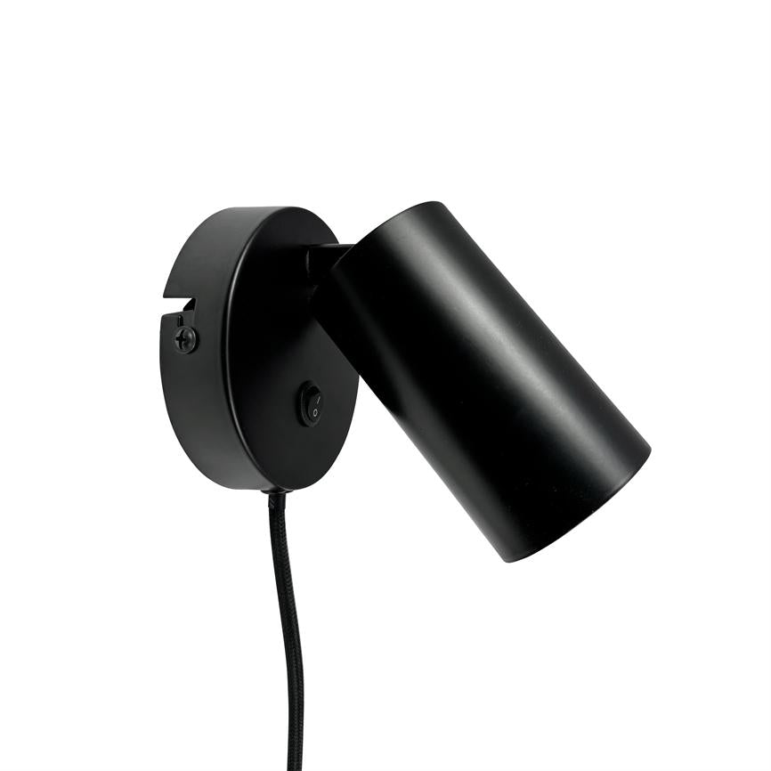 Sort væglampe peger nedad og til højre; mat cylindrisk skærm og rund vægbasis med tænd/sluk-knap, hængende sort tekstilledning mod en ren hvid baggrund.