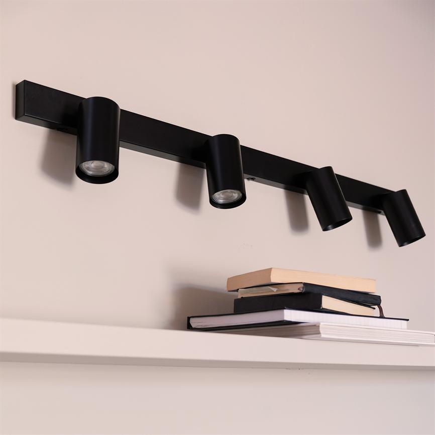 Sort spotlightarm med fire cylindriske lamper monteret på en væg; lamperne er rettet nedad over en hvid hylde, hvor en stak bøger ligger.