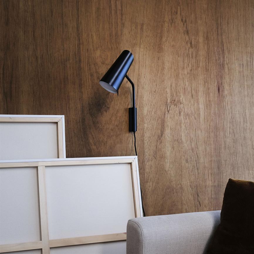 Sort væglampe monteret i en bøjet arm, vendt nedad mod en træbeklædt væg; to blanke lærreder læner sig mod væggen, med en beige sofa og mørk pude i forgrunden.