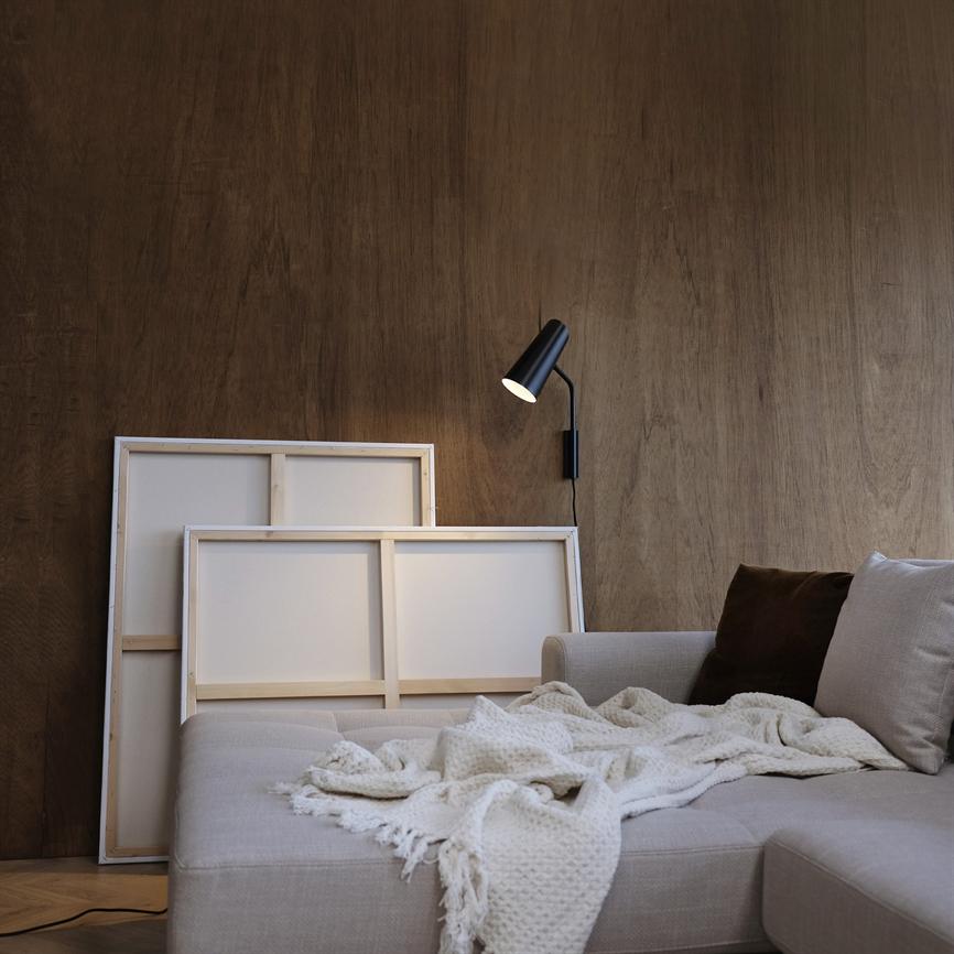 Sofaen er dækket af et krøllet, lyst strikket tæppe; to stående lærreder står bagved, mens en tændt sort væglampe kaster varmt lys på en træbeklædt væg.