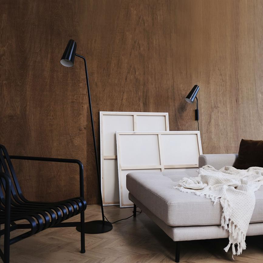 Beige chaiselong dækket af hvidt strikket tæppe står foran to tomme lærreder lænet mod en varm træbeklædt væg, med sort gulvlampe og væglampe samt sort stålstol ved siden.