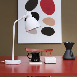 Hvid skrivebordslampe peger ned mod en sort krus på en rød bordplade, flankeret af en hvid boks og en sort kaffetragt; rød stol og abstrakt maleri i baggrunden.