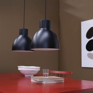 To sorte kuppelformede hængelamper hænger over et rødt bord med en bogstak og et glas; rummet har brune vægge, en rød stol og en hvid skål.